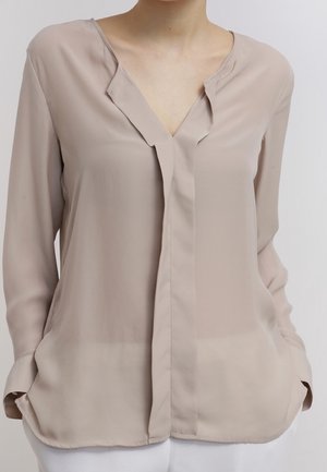 Beige blouse met lange mouwen, een V-hals en een verborgen knoppenlijst, gedragen met witte broek, handen in de zakken gestoken.
