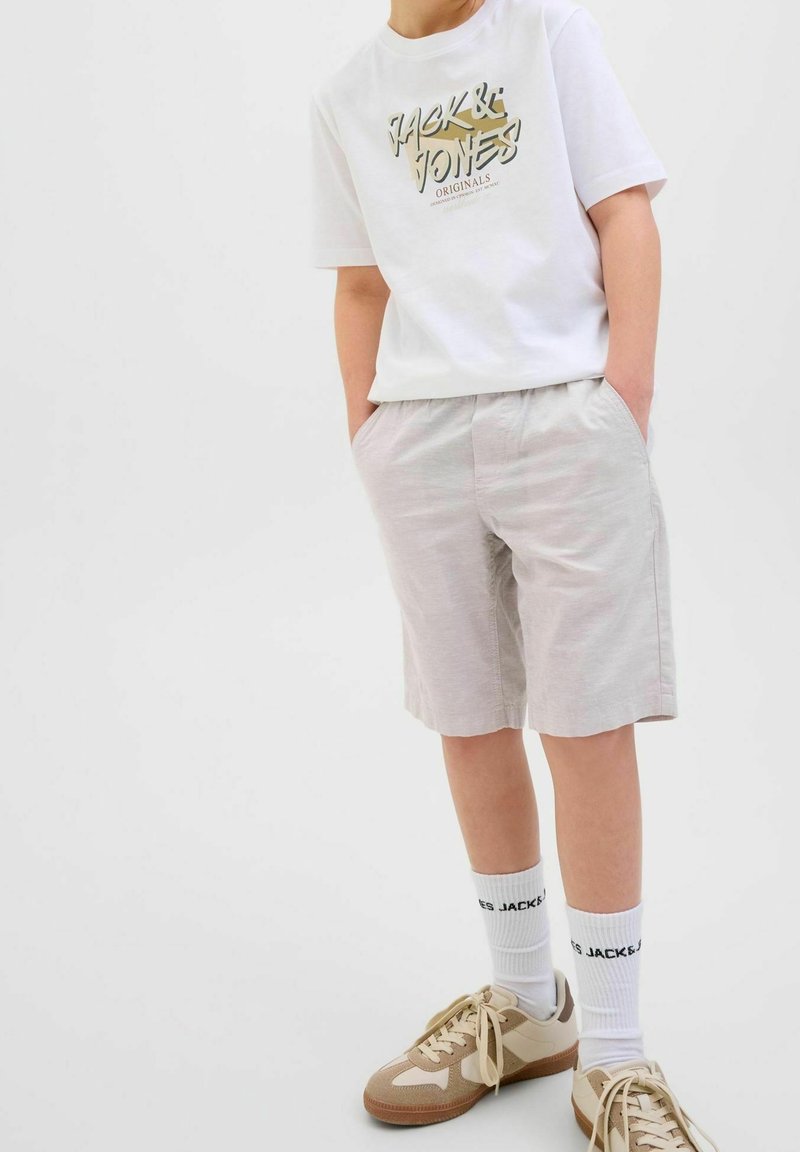 Enfant portant un T-shirt blanc à motif, un short beige clair, des chaussettes blanches avec texte et des baskets beiges, mains dans les poches, debout sur fond blanc.