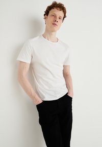 Tânăr cu păr roșcat și creț, purtând un tricou alb simplu cu mâneci scurte și pantaloni negri, stând cu mâinile în buzunare lângă un perete alb.