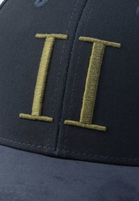 Tumši zila auduma cepure ar plakanu vizieri; priekšpusē izšūti biezā un zelta krāsā "II" burti; gluda tekstūra ar pastiprinātu šuvju apdari.