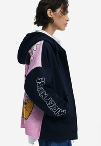 Marineblauer Hoodie mit einem pinken Strickbereich und Cartoon-Grafiken. Weißer Text auf dem Ärmel lautet "Mickey Mouse." Weiches, lässiges Material.