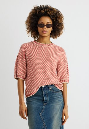 Γυναίκα με σγουρά μαλλιά φορώντας σκούρα γυαλιά ηλίου, ένα ροζ πουλόβερ με κοντά μανίκια και σχέδιο και μια μπλε τζιν φούστα μπροστά σε απλό φόντο.