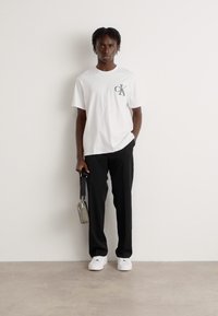 Hvid crew-neck t-shirt med et lille "CK" logo, parret med sorte bukser og hvide sneakers. Personen holder en sølvfarvet taske.