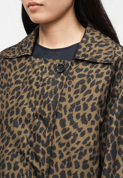 Γυναίκα που φοράει παλτό με μοτίβο λεοπάρ σε σκούρο ελιάς και μαύρο, με γιακά και ένα μαύρο κουμπί, πάνω από μαύρο τοπ.