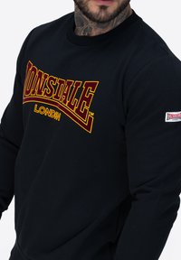 Sudadera negra con un llamativo logo rojo y amarillo de "Lonsdale London", cuello redondo y ribeteado. Material de algodón suave, corte ajustado.