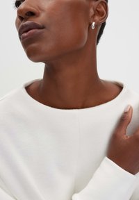 Maglione bianco, strutturato, con scollatura ampia. Primo piano di una spalla di una persona, che mette in risalto la pelle liscia e un piccolo orecchino d'argento.