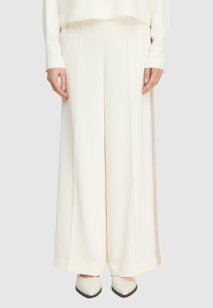 Kobieta ubrana w kremowe, szerokie spodnie z wysokim stanem, z plisami z przodu oraz pasujące szpiczaste buty, ręce luźno opuszczone wzdłuż ciała.