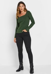 Grünes langärmliges Oberteil mit quadratischer Ausschnitt, figurbetontem Schnitt und weichem Material, kombiniert mit schwarzen Skinny-Jeans und Ankle Boots.