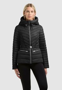 Donna che indossa una giacca imbottita nera trapuntata con cappuccio, cintura in vita e tasche con zip, in piedi contro uno sfondo chiaro uniforme.