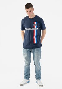 Navy blue cotton t-shirt featuring white and red vertical stripes, logo text “oasis” and “knebworth” across the front. Standard fit.