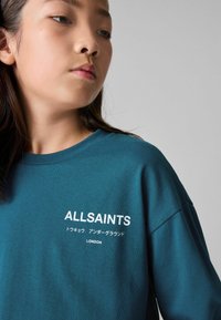 Teal pamučna majica s okruglim izrezom, na kojoj se s lijeve strane prsiju nalazi bijeli natpis "ALLSAINTS" i japanski znakovi.