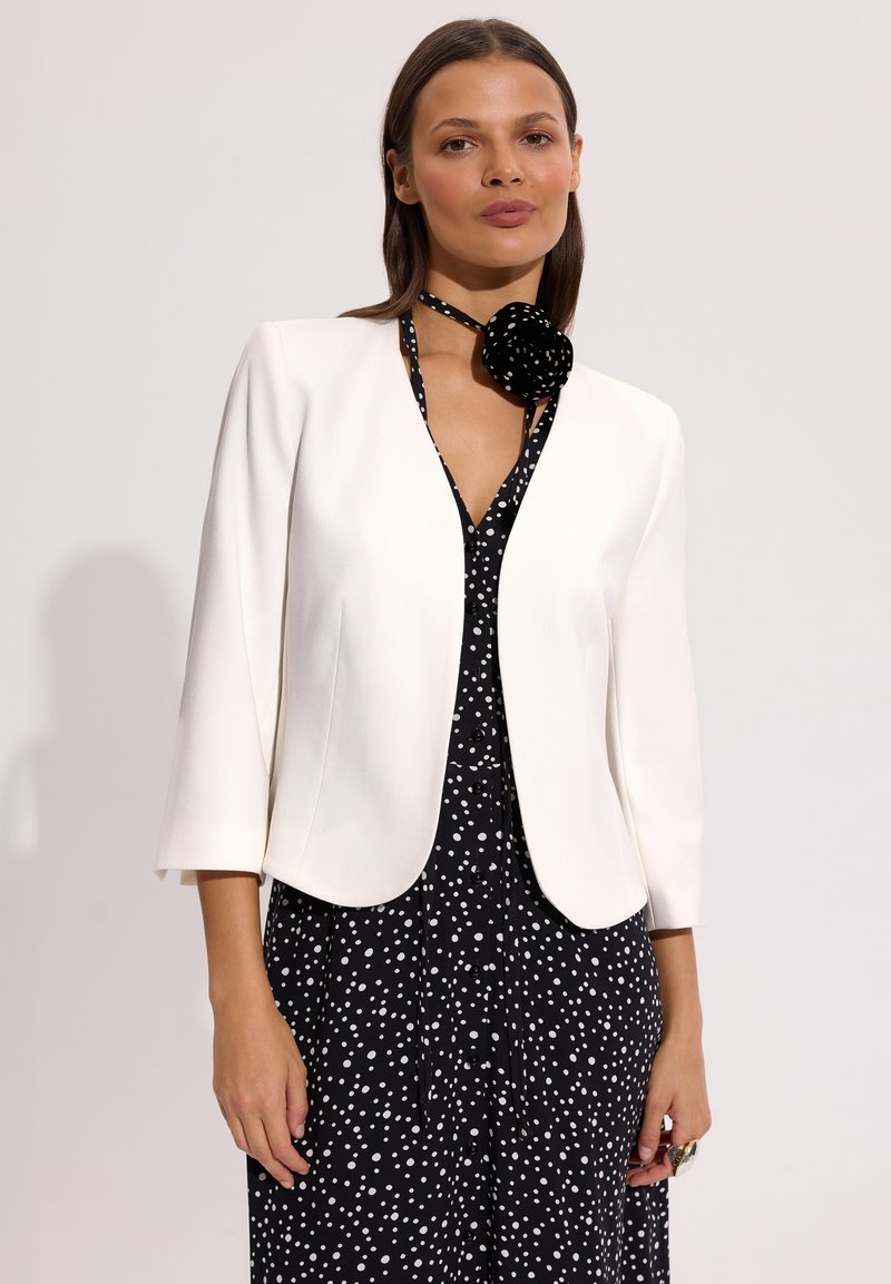 Kobieta ubrana w biały, otwarty blezer na czarnej sukience w białe kropki oraz dopasowaną apaszkę w groszki na szyi.