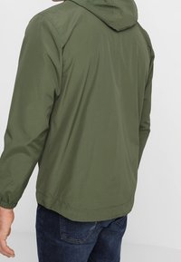 Homme portant une veste à capuche olive avec des poignets élastiques et un jean bleu foncé, vu de dos sur un fond uni.