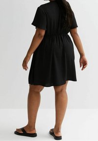 Black short-sleeve dress with an elastic waist, flared skirt, and a smooth texture. Worn with black flat sandals, viewed from the back.
