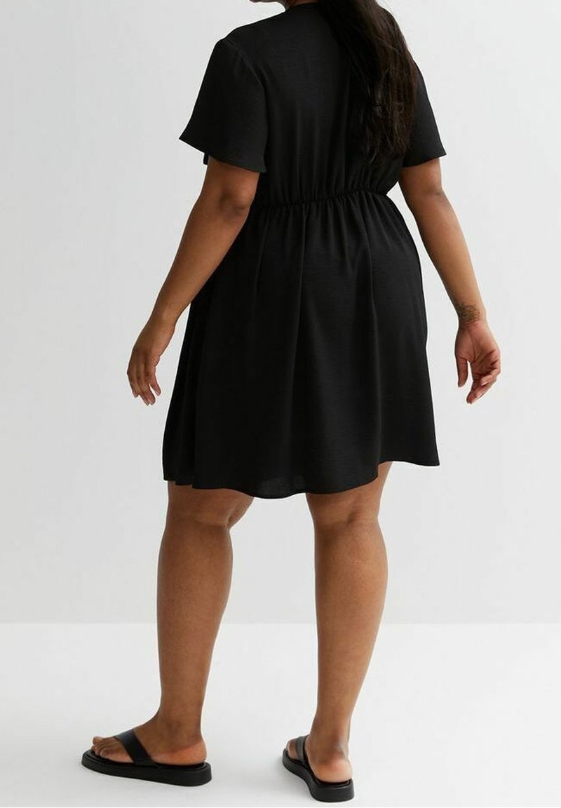 Black short-sleeve dress with an elastic waist, flared skirt, and a smooth texture. Worn with black flat sandals, viewed from the back.
