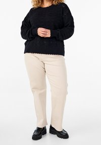 Person, der einen schwarzen strukturierten Pullover, beige Hose und schwarze klobige Loafer trägt und mit locker verschränkten Händen dasteht.