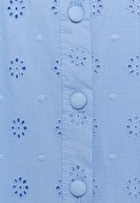 Tessuto di cotone azzurro chiaro con ricami floreali, caratterizzato da bottoni rotondi e dettagli testurizzati. Diversi piccoli intagli formano un motivo.