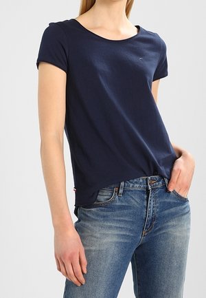 Vrouw die een donkerblauw T-shirt met korte mouwen en een blauwe spijkerbroek draagt, met één hand in de zak en staande tegen een effen achtergrond.