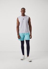 Witte mouwloze shirt met een textuurpatroon, lichtblauwe shorts, marineblauwe legging en witte sneakers. Adidas-logo op shirt en shorts.