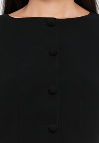 Gros plan d’un vêtement noir avec un col rond et quatre boutons visibles recouverts de tissu noir sur le devant.