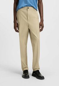 Pantaloni beige con taglio dritto, caratterizzati da una piega frontale singola e chiusura con bottone, abbinati a scarpe nere.