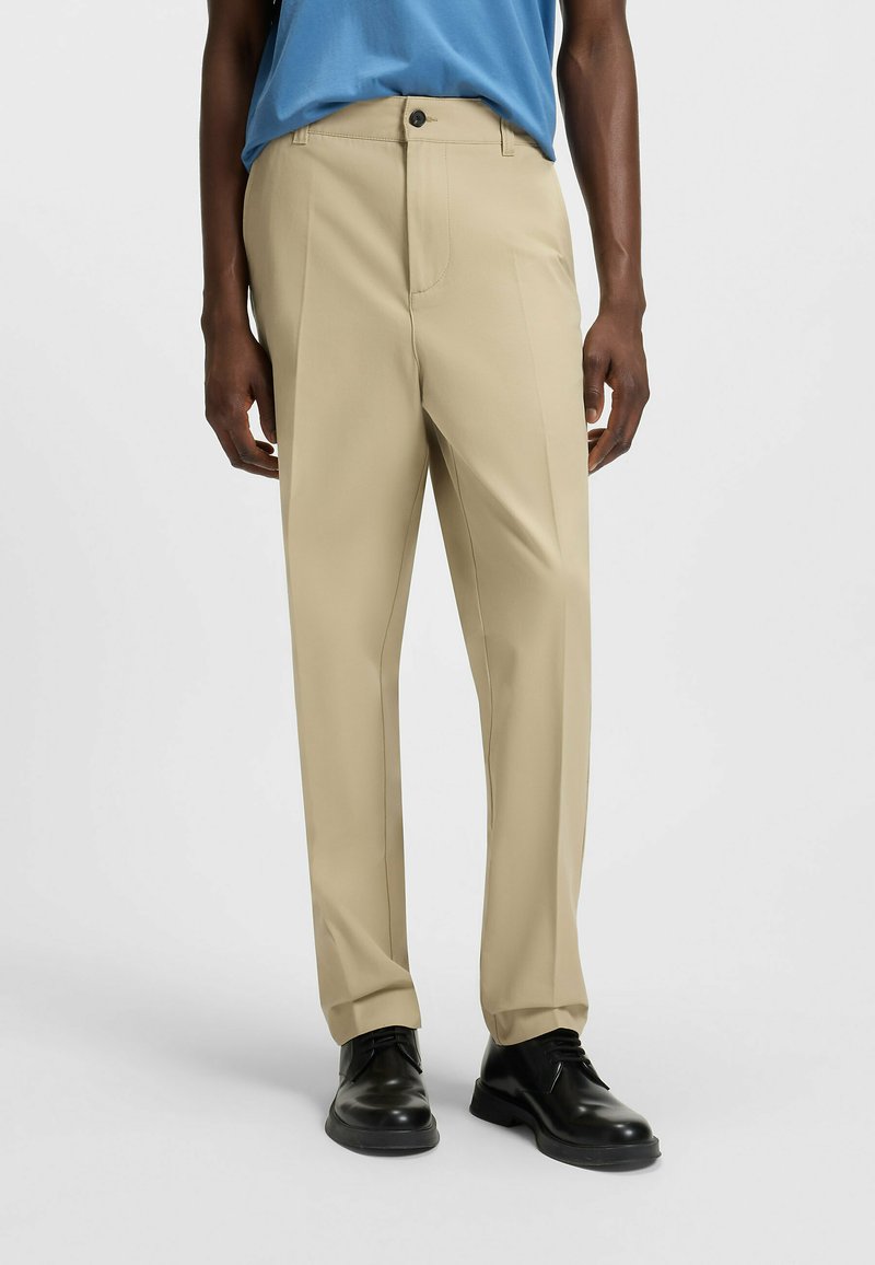Pantaloni beige con taglio dritto, caratterizzati da una piega frontale singola e chiusura con bottone, abbinati a scarpe nere.