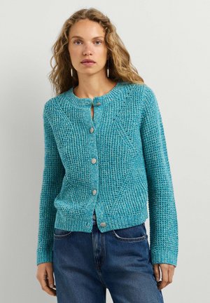 Vrouw draagt een blauwe, getextureerde gebreide cardigan met knopen en blauwe spijkerbroek, staand tegen een effen lichte achtergrond.