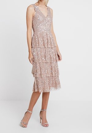 Bezruké šaty délky po kolena v růžově zlaté barvě s flitry a vrstvenými volány, nošené stojící ženou se stříbrnými sandály na vysokém podpatku.