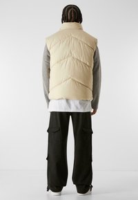 Chaleco de plumas en beige claro con costuras en zigzag, llevado sobre un suéter gris, combinado con pantalones cargo negros holgados, que cuentan con bolsillos laterales.