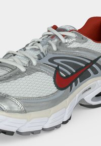 Γκρι και λευκό αθλητικό παπούτσι Nike με κόκκινο σήμα, λευκά κορδόνια, σόλα Cushlon και κόκκινη εσωτερική επένδυση, εμφανίζεται με γωνία από το πλάι.