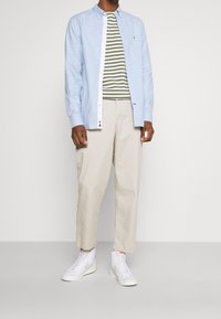 Lichtblauw overhemd over een groen-wit gestreept T-shirt, gecombineerd met beige broek en witte sneakers.