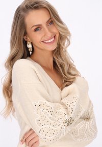 Beige kötött pulóver hosszú ujjal, átlátszó virágmintákkal díszítve, amelyeket flitterek és gyöngyök ékesítenek, texturált mintát létrehozva.
