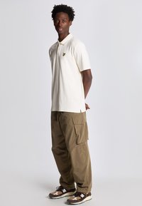 Polo shirt in crème met een logo op de voorkant, gedragen met losse olijfgroene cargo-broeken met zijzakken en beige-brown sneakers.