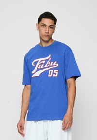 Blå kortermet T-skjorte i bomull, med et stort hvitt og rødt "Fubu 05"-logo på forsiden. Enkel design med crew neck.