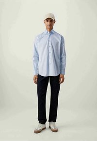 Camisa de mangas compridas azul clara com botões, calças azul marinho e boné bege. Apresenta ténis casuais com parte superior branca e solas bejes.