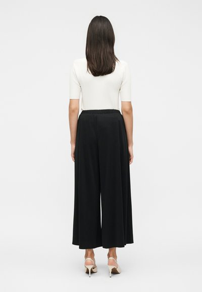 Γυναίκα στέκεται με την πλάτη προς τα εμπρός, φοράει λευκό κοντομάνικο τοπ, μαύρο παντελόνι με φαρδιά πόδια και μπεζ σανδάλια με ψηλό τακούνι σε μονόχρωμο φόντο.