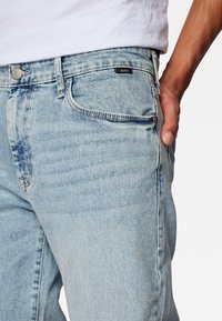 Jeans de mezclilla azul claro con un diseño clásico de cinco bolsillos, un ligero desvanecimiento y una etiqueta de la marca Mavi en la cinturilla.