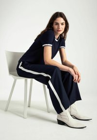 Marineblaue T-Shirt mit weißem Saum, kombiniert mit weit geschnittenen marineblauen Hosen mit weißen Seitenstreifen und weißen spitzen Ankle Boots.