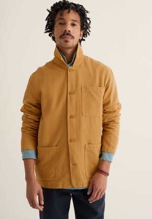 Uomo con capelli ricci che indossa una giacca giallo senape con bottoni sopra una camicia azzurra, pantaloni scuri e un braccialetto intrecciato al polso destro.