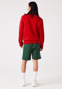 Sweat-shirt rouge avec poignets côtelés, associé à un short vert avec des accents latéraux bleu clair. Des baskets blanches et des chaussettes complètent le look.