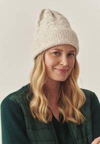 Vaaleanbeige neulottu myssy, jossa on teksturoitu pinta ja käännetty reuna, jonka päällä on pitkä, laineikasta hiusta ja vihreä takki.