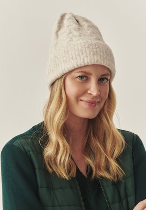 Svetlo béžová pletená čapica s textúrovaným povrchom a zavinutým okrajom, ktorý nosí osoba s dlhými, vlnitými vlasmi a zelenou bundou.