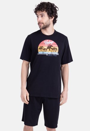 Homme aux cheveux bouclés portant un short noir et un T-shirt noir arborant un motif coloré de coucher de soleil et de palmier avec le texte "Vintage Athena".