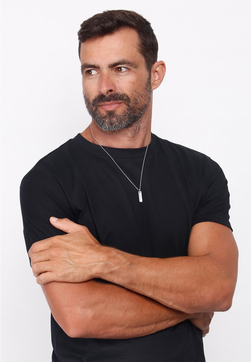 Sort kortærmet skjorte båret af en mand med en sølvvedhæng. Vedhænget er cylindrisk med et let vertikalt design.