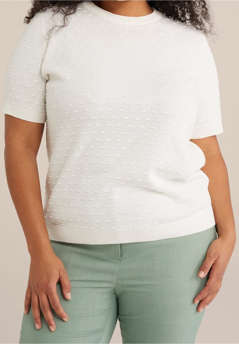 Biały sweter z krótkim rękawem z teksturowanymi kropkami, okrągłym dekoltem i prążkowanym dołem. Połączony z jasnymi zielonymi spodniami.
