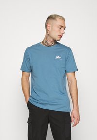 Blaues Baumwoll-T-Shirt mit Rundhalsausschnitt und kurzen Ärmeln. Mit kleinem weißen Logo auf der linken Brust. Glatte Textur.
