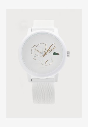 Montre-bracelet blanche avec bracelet en silicone texturé, initiales dorées en écriture cursive sur le cadran et petit logo crocodile vert Lacoste à la position trois heures.