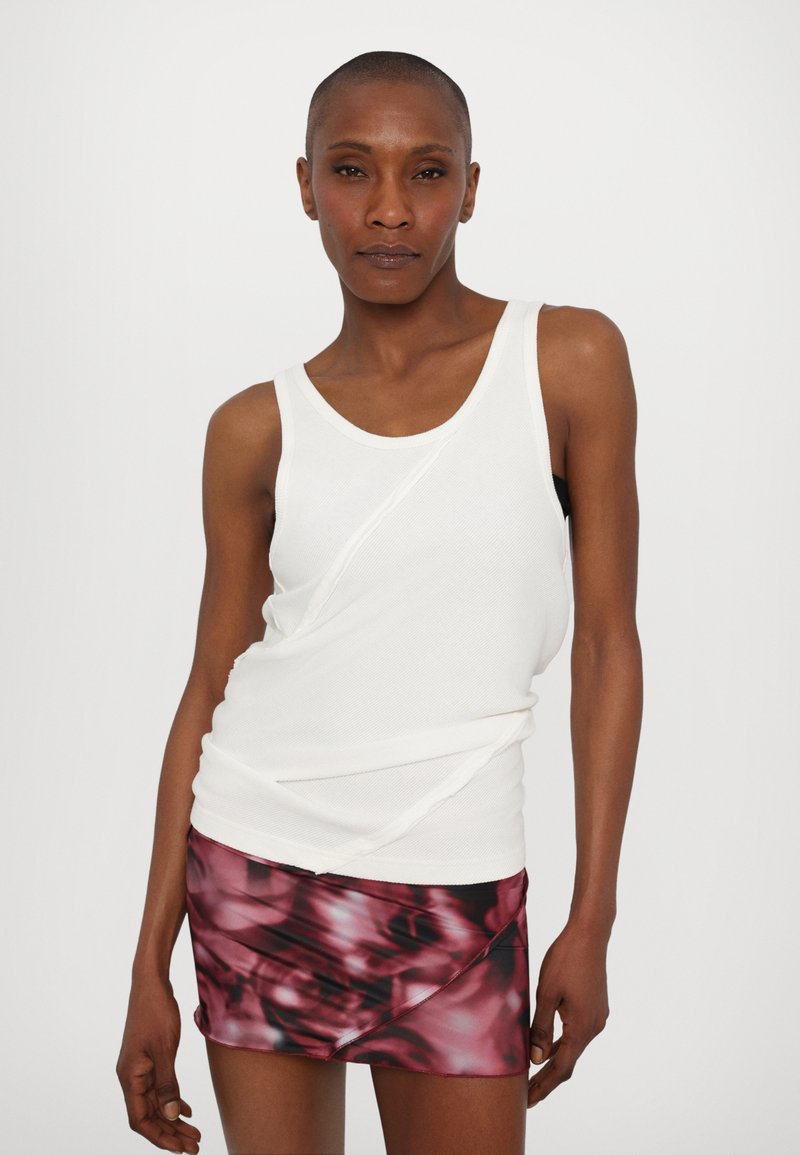 Witte mouwloze top met ribbeltextuur, voorzien van een asymmetrisch ontwerp. Gecombineerd met een korte, donkerroze met een patroon.