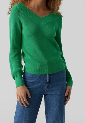 Mujer con un suéter de color verde brillante con cuello en V, mangas largas y jeans azules, de pie contra un fondo claro y liso.