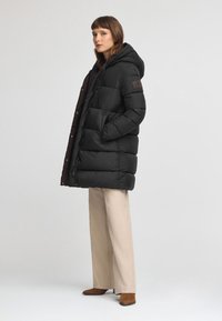 Czarna kurtka puchowa z kapturem, z pikowanym wzorem, bocznymi kieszeniami i naszywką z logo na rękawie; w połączeniu z kremowymi spodniami.